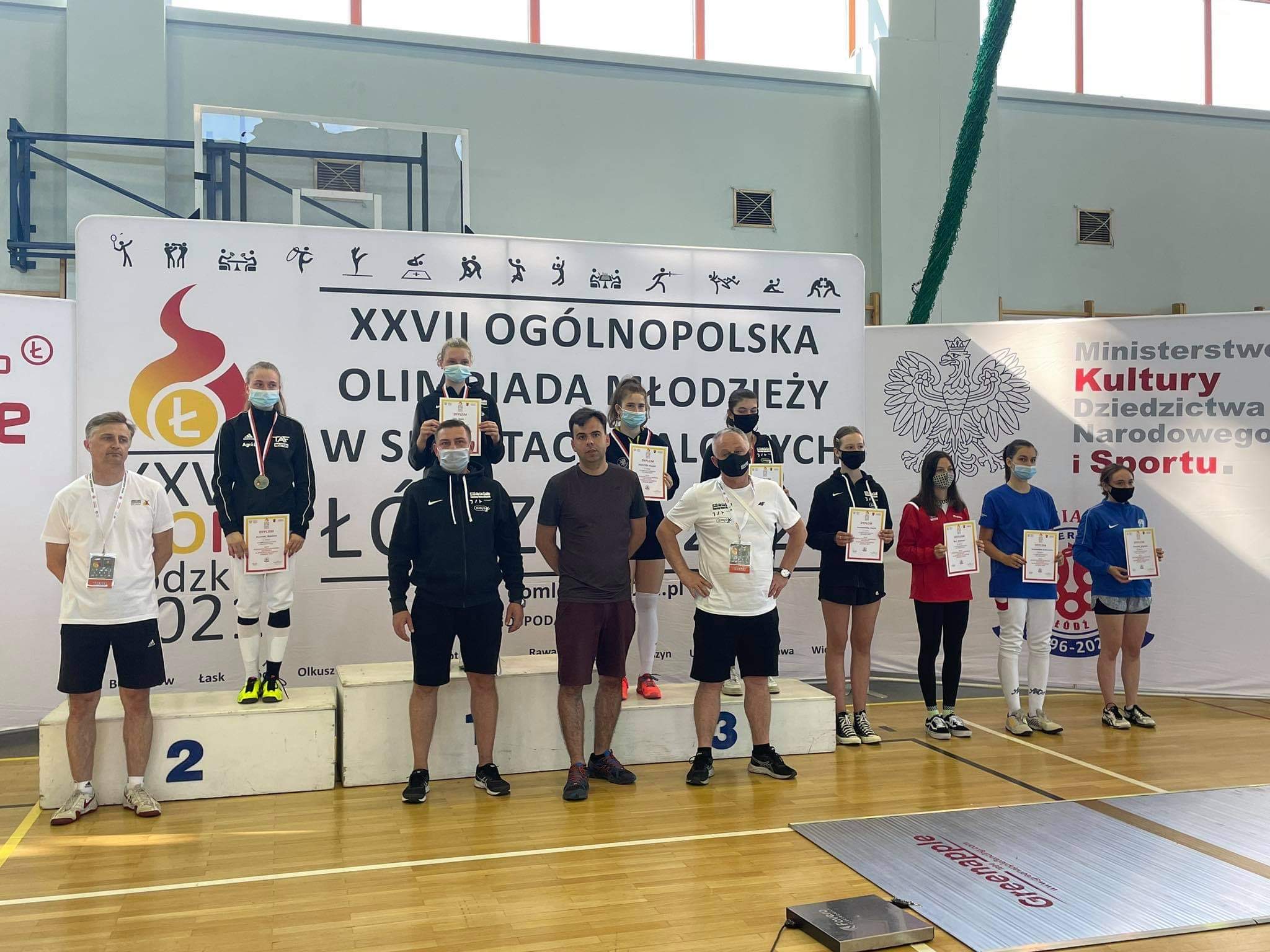 Zdjęcie zawodników na podium oraz trenerów stojących przed podium.