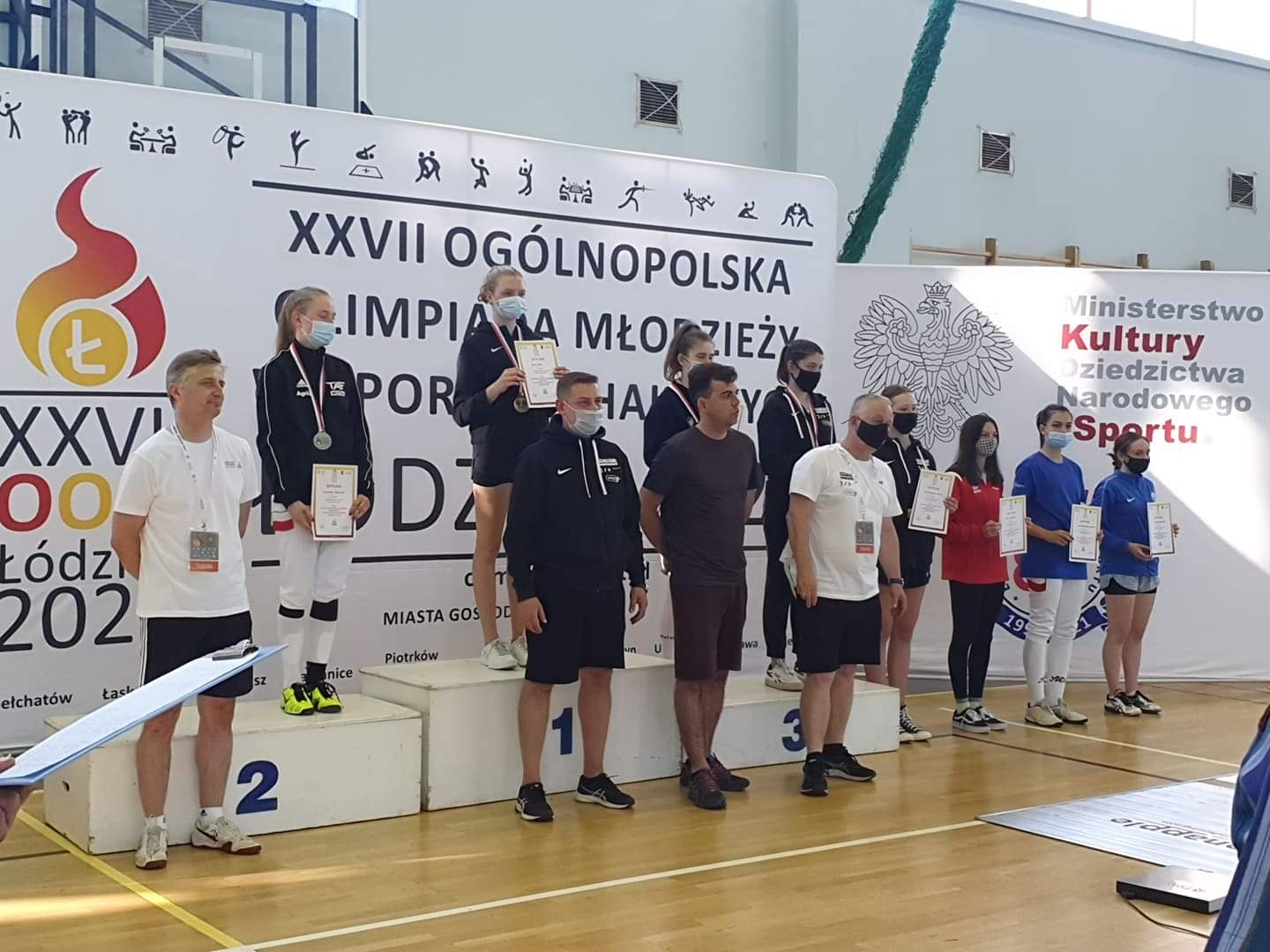 Zdjęcie zawodników na podium oraz trenerów stojących przed podium.